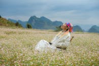 Woman in colorful headscarf relaxes on chair in vibrant flower field with scenic mountain background.