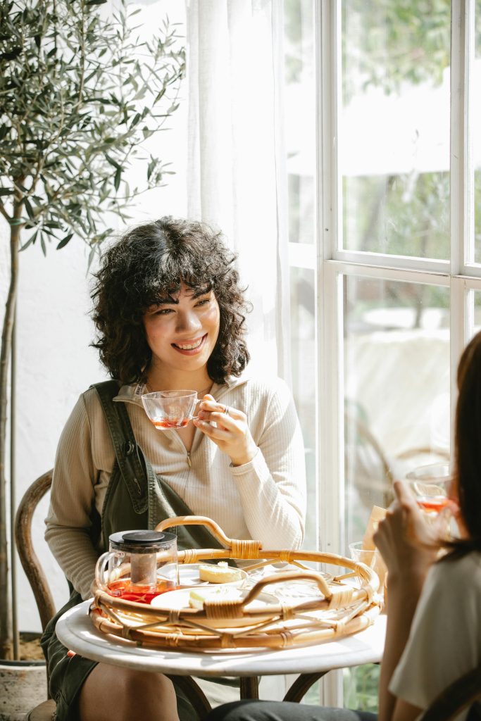 Smiling ethnic female with curly hair sitting at table with breakfast and hot natural tea and talking to girlfriend