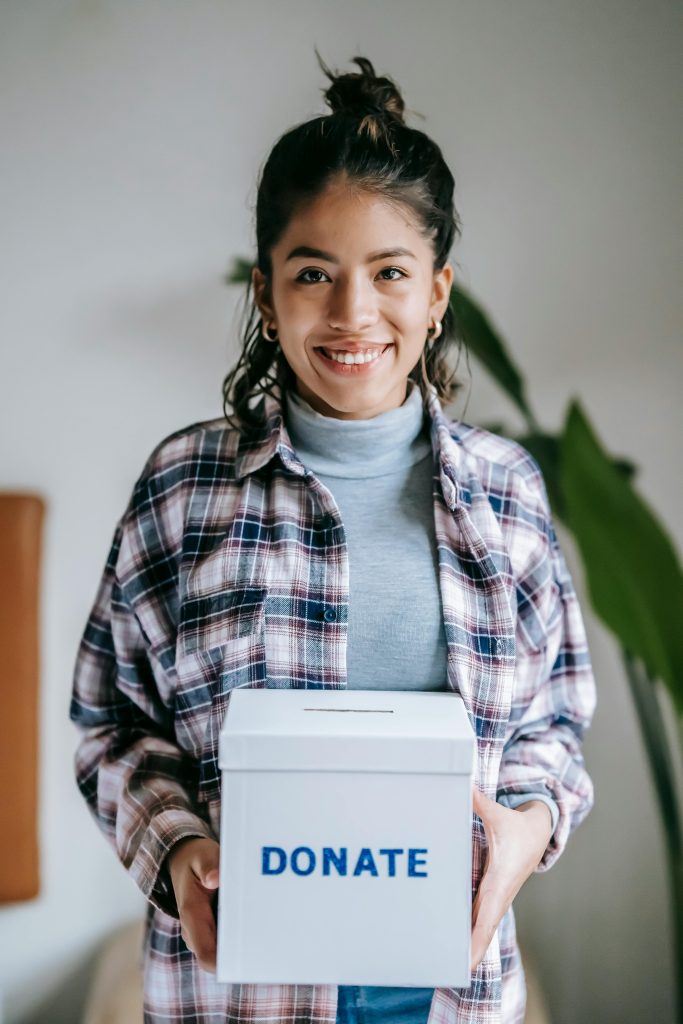 Positive young ethnic female volunteer in checkered shirt with box with inscription donate smiling and looking at camera