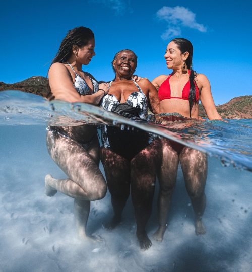 Three women laughing together in the clear ocean water on a sunny day, captured from a unique perspective.