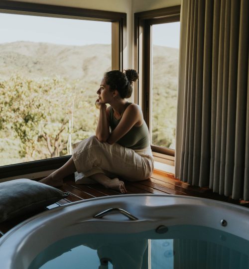Woman gazing through window beside indoor jacuzzi overlooking mountains.