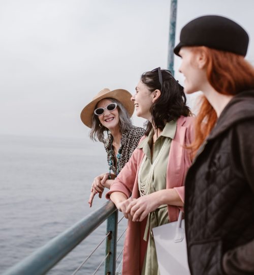 Three women laughing together, enjoying a sunny day by the sea, showcasing friendship and travel.