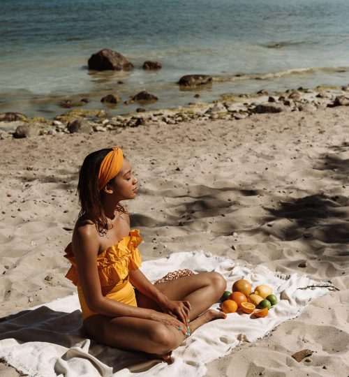 Serene moment of a woman in an orange swimsuit sitting on a beach alongside tropical fruits.
